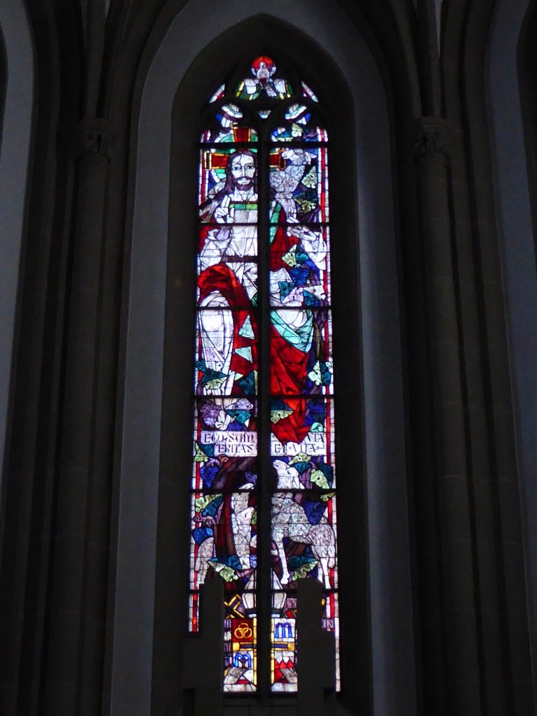 Chorfenster Mitte: Zu sehen ist Jesus als Guter Hirte und der lateinische Schrifttext: „Ich bin der Weg, die Wahrheit und das Leben.“ (Joh 14) Darunter findet sich das Wappen von Julius Döpfner (Bischof von Würzburg 1948-1957) mit seinem Wahlspruch: „Wir aber verkündigen Christus, den Gekreuzigten.“
