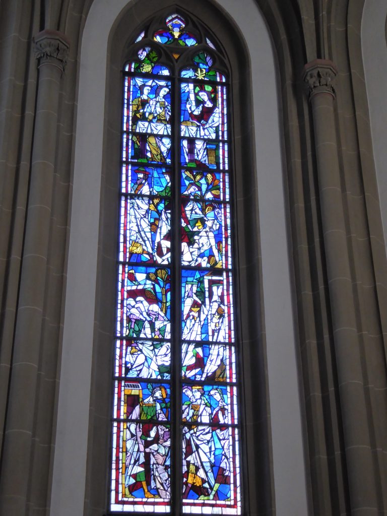 Linkes Chorfenster: Das linke Fenster steht für das Neue Testament. Von unten nach oben: Das Gleichnis von den klugen Jungfrauen (Mt 25), das Gleichnis vom verlorenen Sohn (Lk 15), das Gleichnis vom Barmherzigen Samariter (Lk 10) und die Begegnung des Auferstandenen mit den Emmausjüngern (Lk 24).